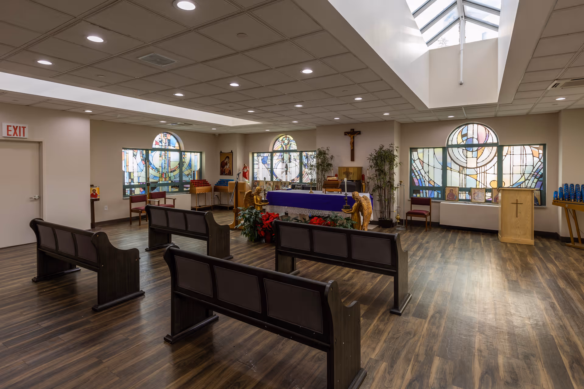 Interior view of a chapel with wooden pews, stained glass windows, religious statues, and a crucifix on the wall. The room has a wooden floor, a skylight, and various religious icons and decorations.