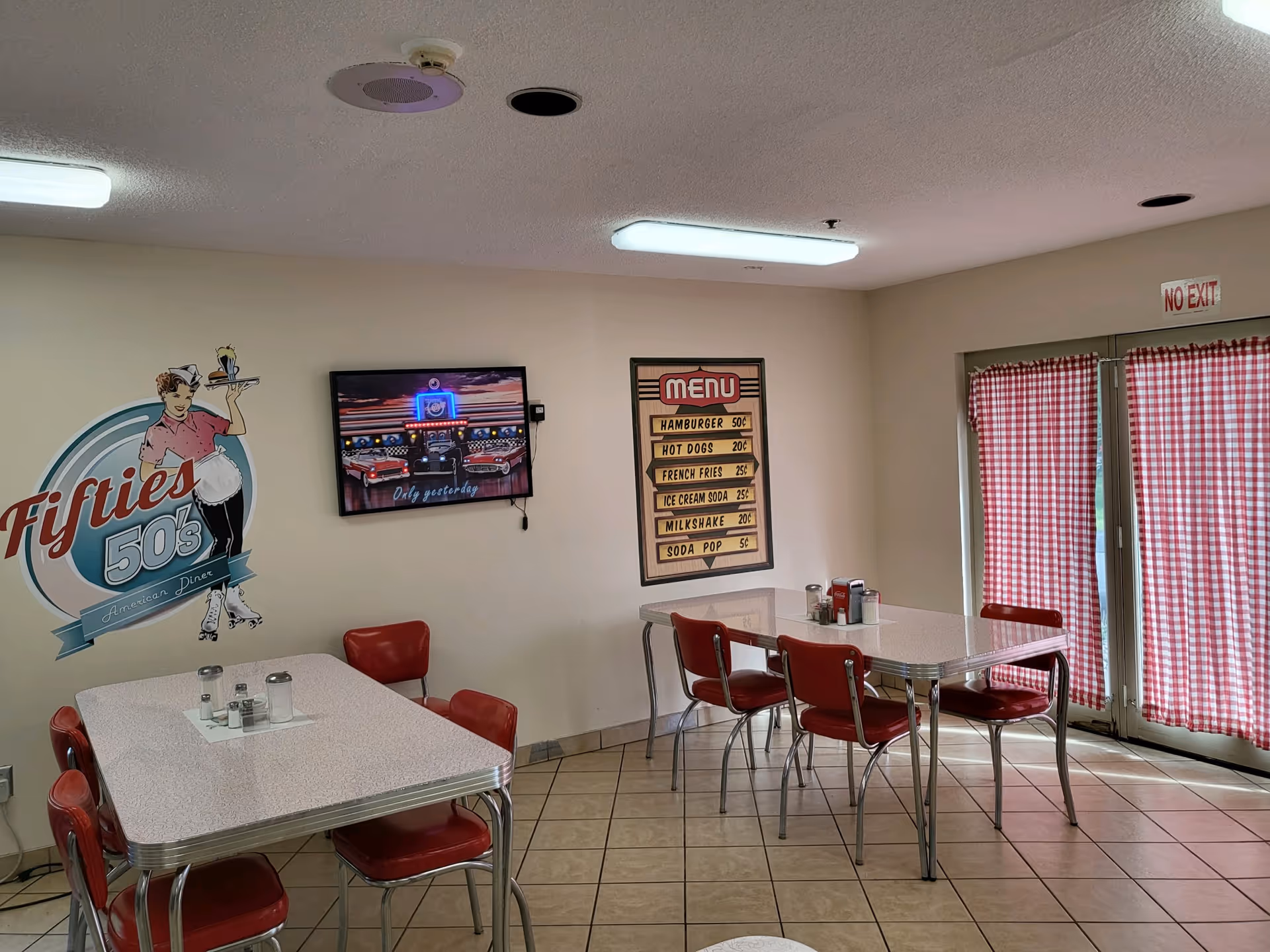 Small 1950s-style diner dining area with red chairs, retro wall decor and checkered curtains.