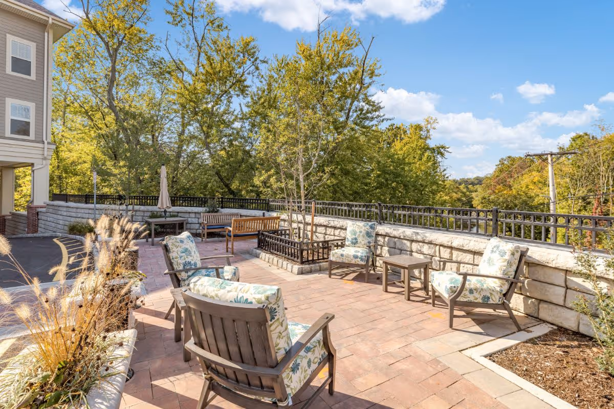 Outdoor patio area at Sunrise of Summit with cushioned chairs arranged around small tables on a brick-paved surface, surrounded by a stone wall and black metal railing, with trees and blue sky in the background.