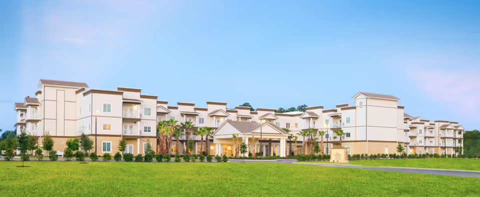 Panoramic front view of a three-story senior living building with a manicured lawn, palm trees, and a covered entrance under a blue sky.
