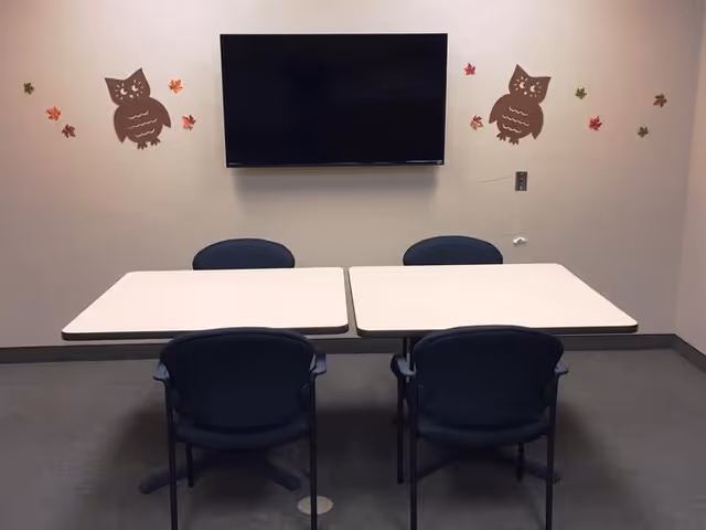 A small meeting or activity room with two white tables pushed together and four black chairs around them. A flat screen TV is mounted on the beige wall above the tables. The wall is decorated with two brown owl decals and several colorful autumn leaves.