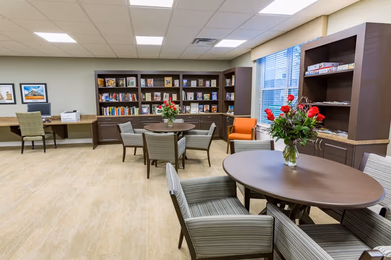 A well-lit room with round tables and striped chairs arranged around them. Each table has a vase with red flowers. There are bookshelves filled with books and board games along the walls. A window with blinds lets in natural light, and there is a desk with a computer and printer in the corner.