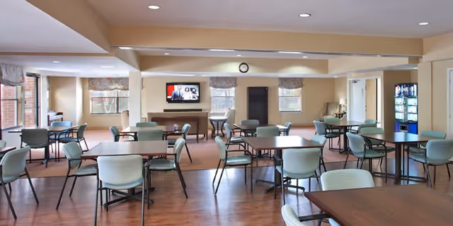 A spacious dining area with multiple square tables and green cushioned chairs arranged neatly. The room has large windows with floral valances allowing natural light to enter. A television is mounted on the far wall above a piano, and a vending machine is visible on the right side. The flooring is a combination of wood and carpet, and the ceiling has recessed lighting.