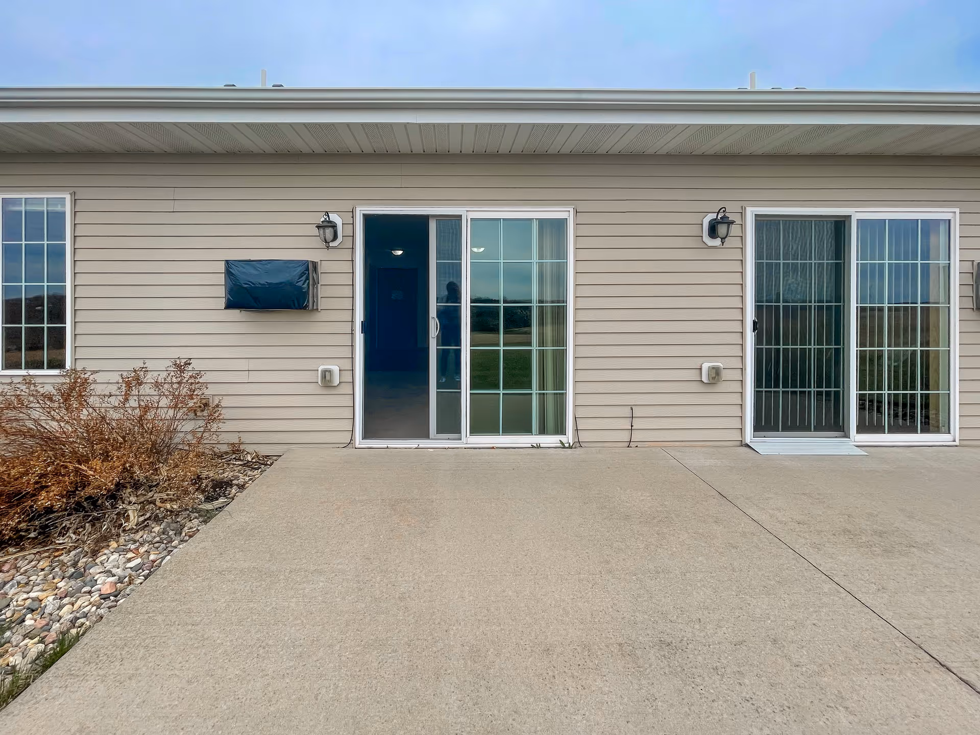 Exterior view of a beige siding building with two sliding glass doors and two wall-mounted light fixtures. There is a concrete patio in front and a small landscaped area with rocks and dried plants to the left.