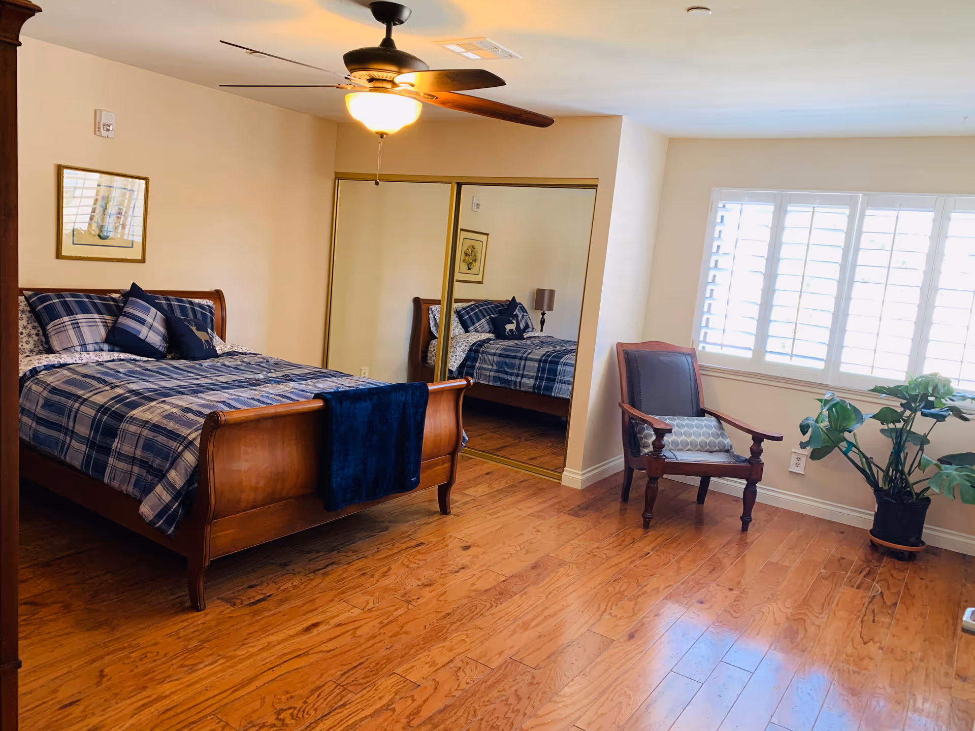 A bedroom with a wooden sleigh bed dressed in blue plaid bedding and multiple pillows. There is a ceiling fan with a light above the bed. A mirrored closet door is on the wall behind the bed. To the right, there is a wooden armchair with a cushion and a potted plant near a window with white shutters. The floor is wooden.
