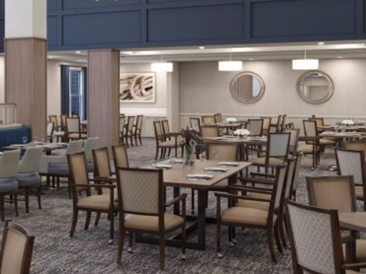 A spacious dining room with multiple wooden tables and cushioned chairs arranged neatly. The room features carpeted flooring, modern pendant lights, two round mirrors on the wall, and a piece of abstract artwork. The setting is elegant and ready for guests with table settings and floral centerpieces.