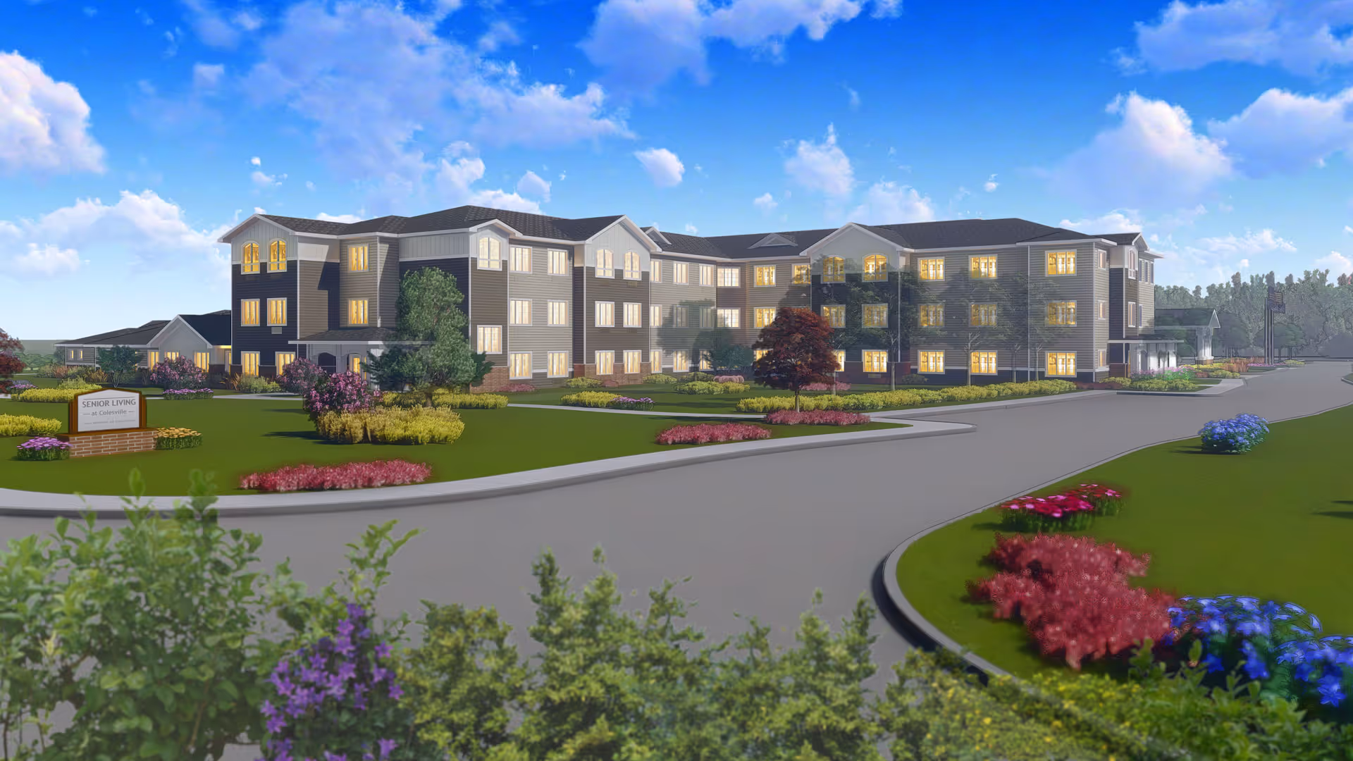 Exterior view of a large three-story senior living facility building with multiple windows lit from inside, surrounded by landscaped gardens with colorful flowers and shrubs under a partly cloudy blue sky.