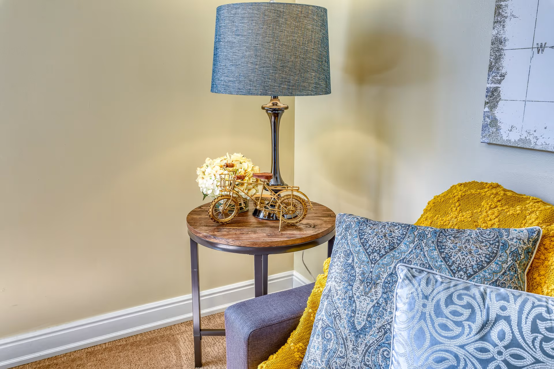 A cozy corner featuring a small round wooden side table with a metal base, holding a decorative lamp with a blue shade, a small golden bicycle sculpture, and a bouquet of white flowers. Next to the table is a gray armchair adorned with a yellow textured throw blanket and two patterned blue and white pillows. The background shows a beige wall with part of a framed artwork visible.