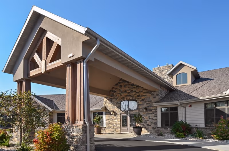 Exterior view of a senior living facility building with a covered entrance supported by wooden beams and stone pillars. The building features stone and siding walls, multiple windows, and a clear blue sky above. There are some shrubs and plants near the entrance.