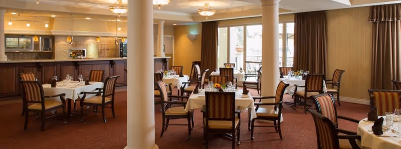 A dining room in a senior living facility with several round tables covered with white tablecloths, each set with glassware, napkins, and small floral centerpieces. The room has carpeted floors, wooden chairs with cushioned seats and backs, large windows with curtains allowing natural light, and a serving counter along one wall with pendant lighting above.