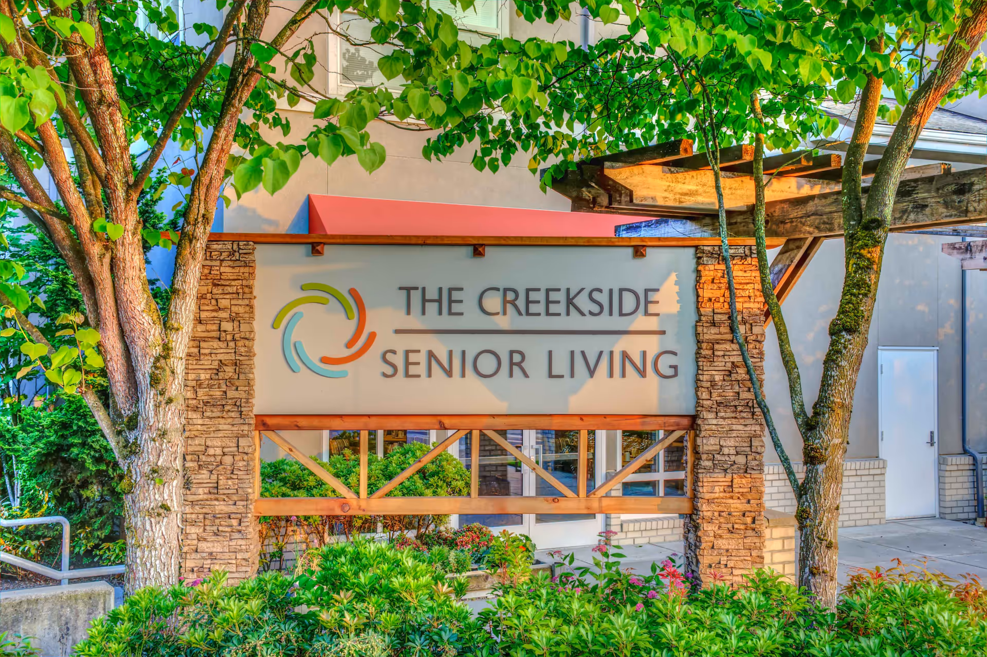 Outdoor sign for The Creekside Senior Living facility surrounded by green trees and shrubs, with a building and a wooden pergola structure in the background.