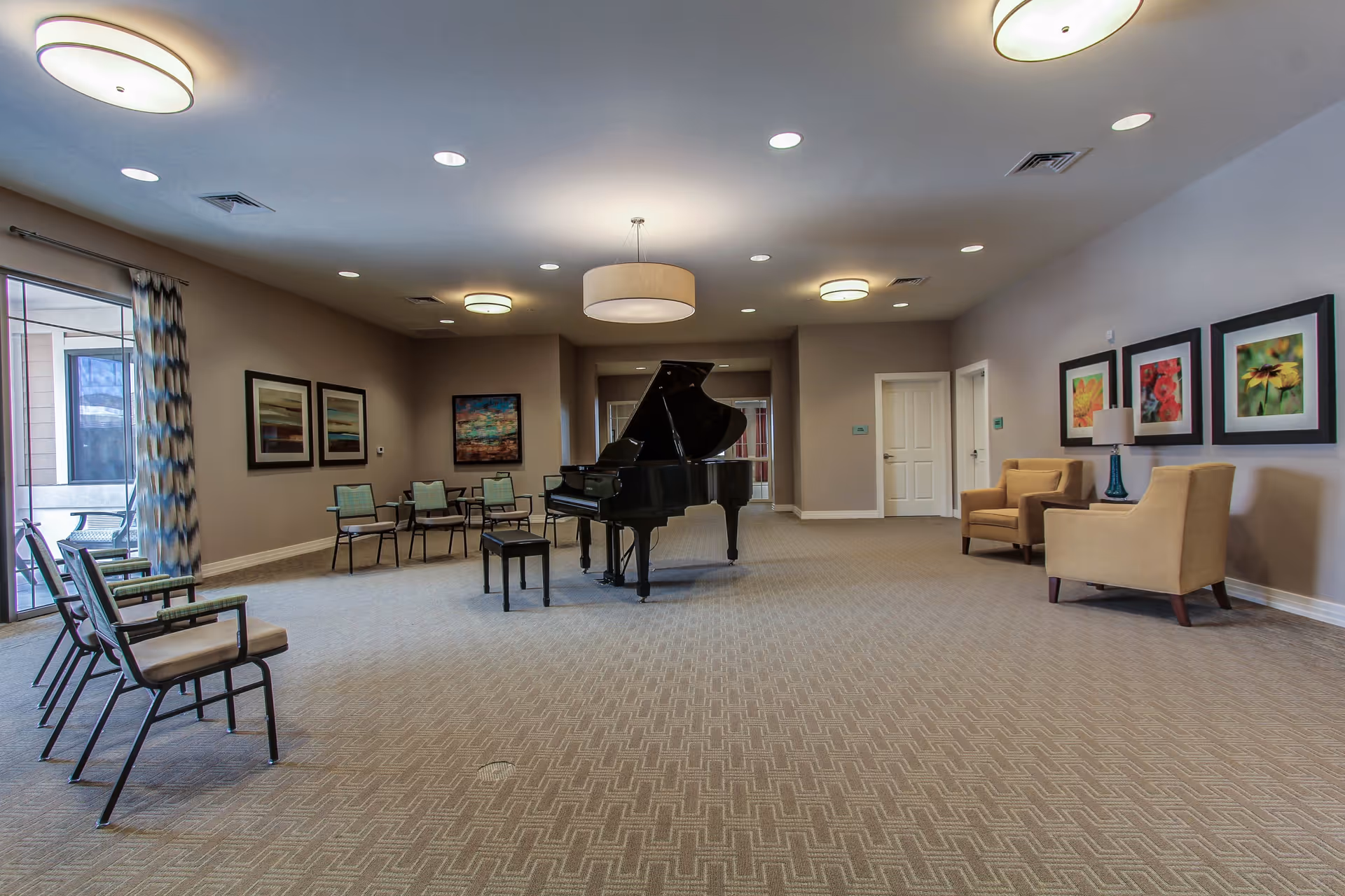 Spacious common room with a grand piano in the center, surrounded by chairs and armchairs. The room has beige walls adorned with framed artwork, carpeted floor, and ceiling lights. Large windows with patterned curtains allow natural light to enter.
