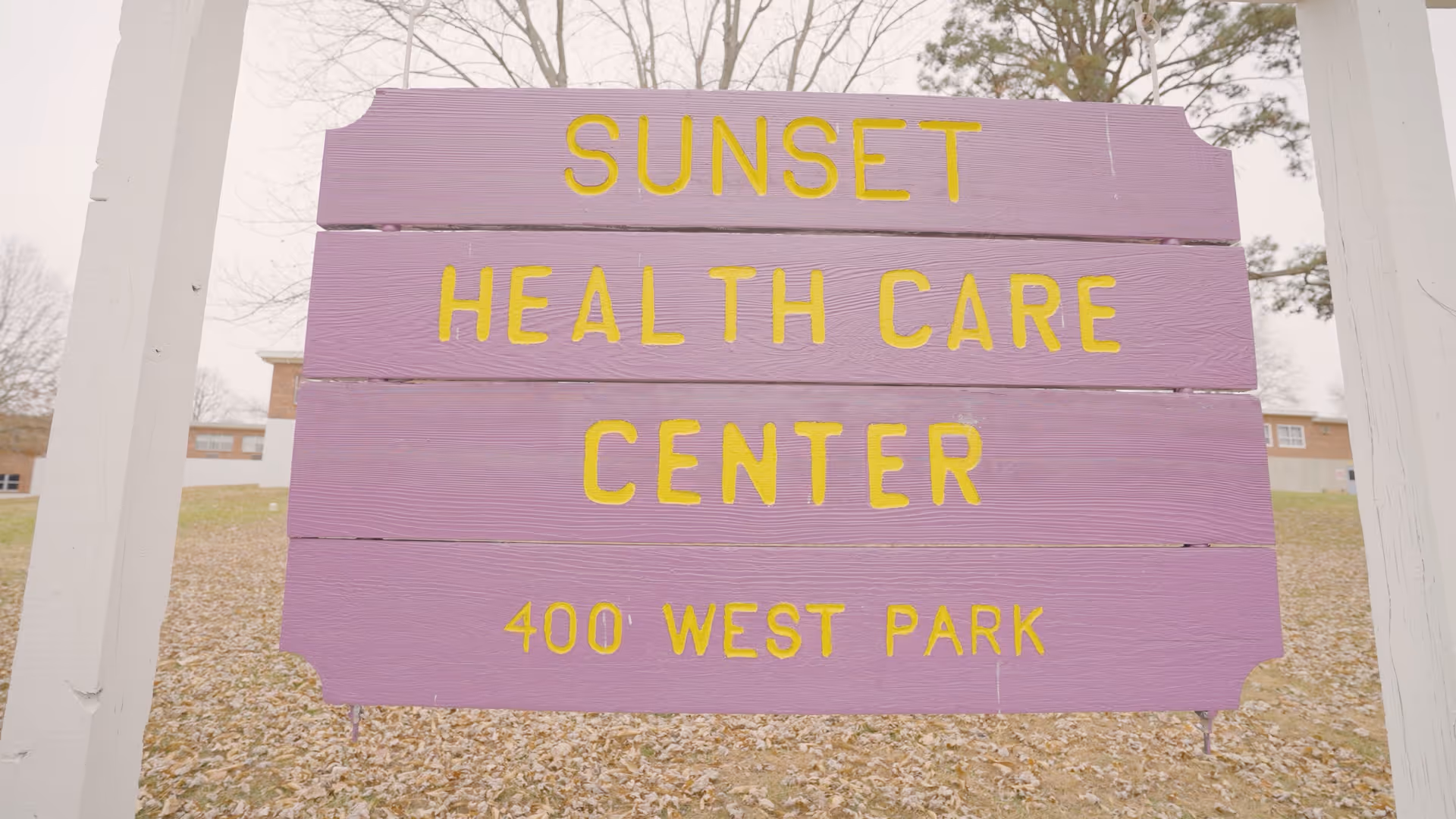 A purple wooden sign with yellow letters that reads 'SUNSET HEALTH CARE CENTER 400 WEST PARK' standing outdoors with a building and leaf-covered ground in the background.