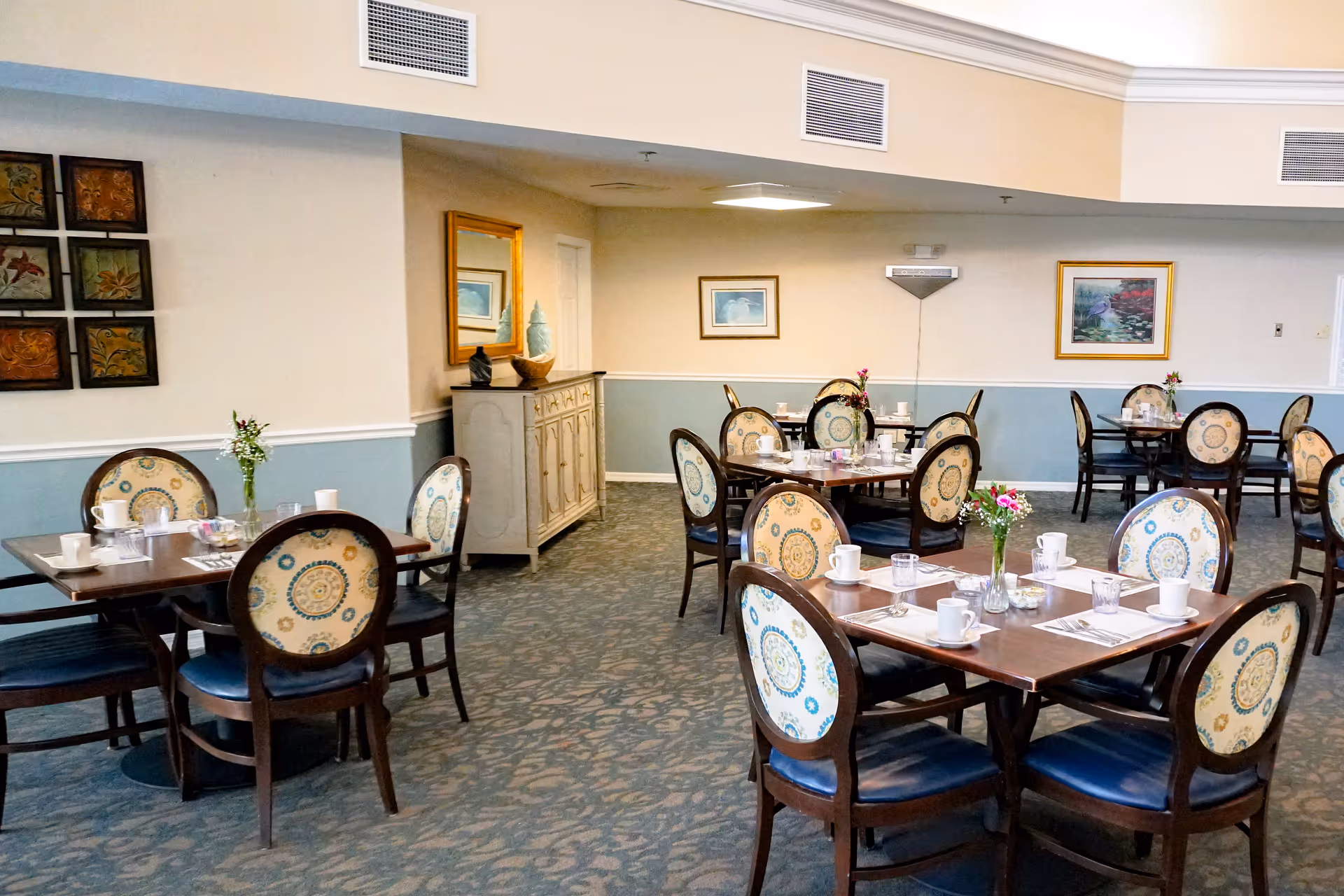 A dining room with several wooden tables and chairs featuring patterned upholstery. Each table is set with white cups, glasses, utensils, and small flower vases. The room has light blue and beige walls adorned with framed artwork and a mirror above a sideboard cabinet.