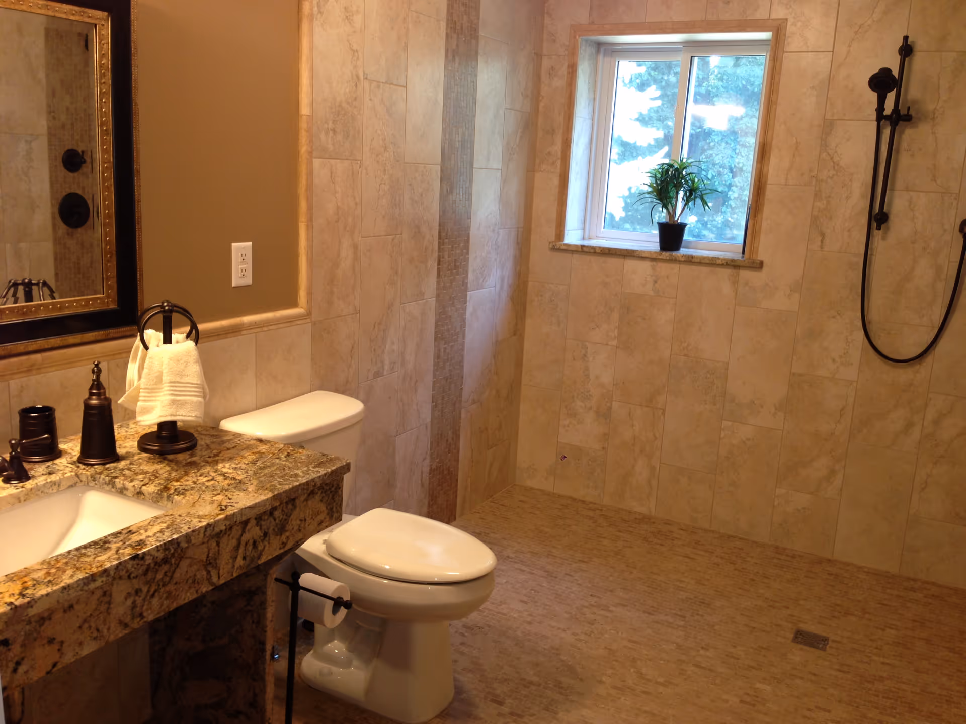A bathroom with beige tiled walls and floor, featuring a white toilet, a granite countertop with a sink, a large mirror, and a window with a small potted plant on the windowsill. There is a black showerhead and handle mounted on the wall to the right.