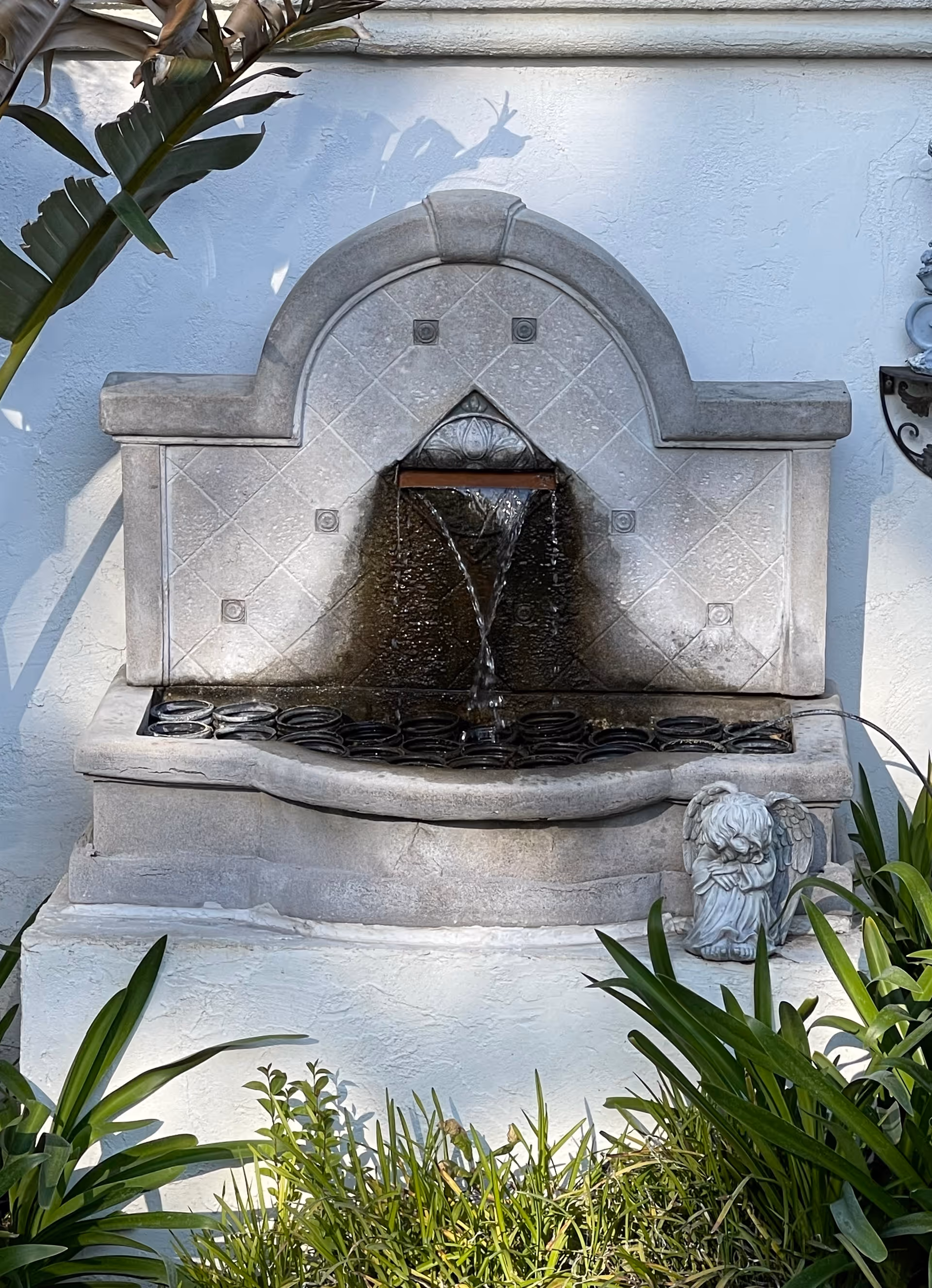 A decorative stone wall fountain with water flowing from a spout into a basin below, surrounded by green plants and a small angel statue on the right side.