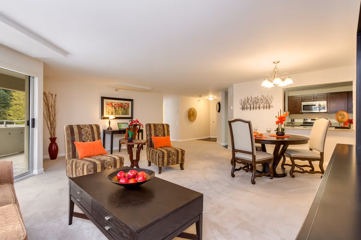 A spacious and well-lit living and dining area in a senior living facility. The room features two patterned armchairs with orange cushions, a dark wooden coffee table with a bowl of red apples, a round dining table with four chairs, and a kitchen area with stainless steel appliances in the background. There is a sliding glass door leading to an outdoor balcony, and the walls are decorated with framed artwork and a metal tree wall sculpture.