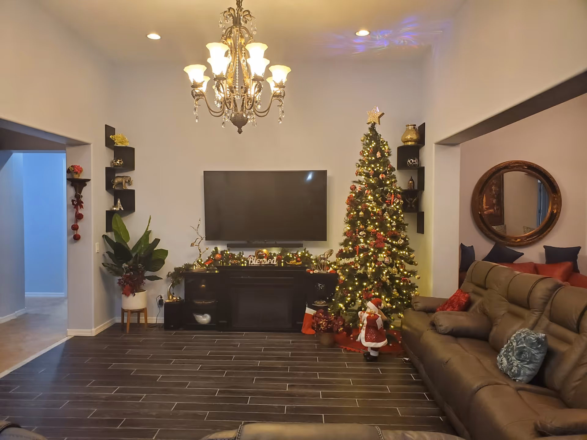 Cozy living room with a decorated Christmas tree, wall-mounted TV, chandelier, and leather sofa.