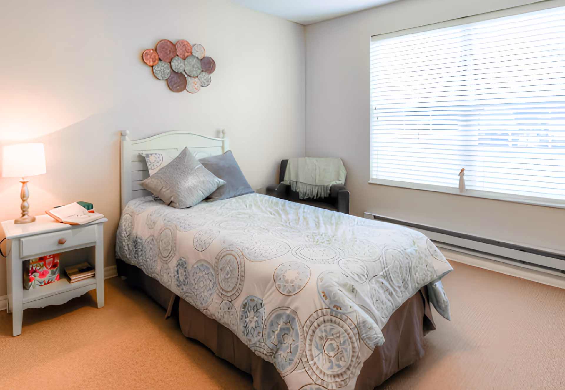 A cozy bedroom with a single bed covered in a patterned quilt and two pillows. Next to the bed is a white nightstand with a lamp, an open book, and some items on the lower shelf. A black armchair with a light-colored throw blanket is positioned near a large window with white blinds. A decorative wall art piece hangs above the bed.