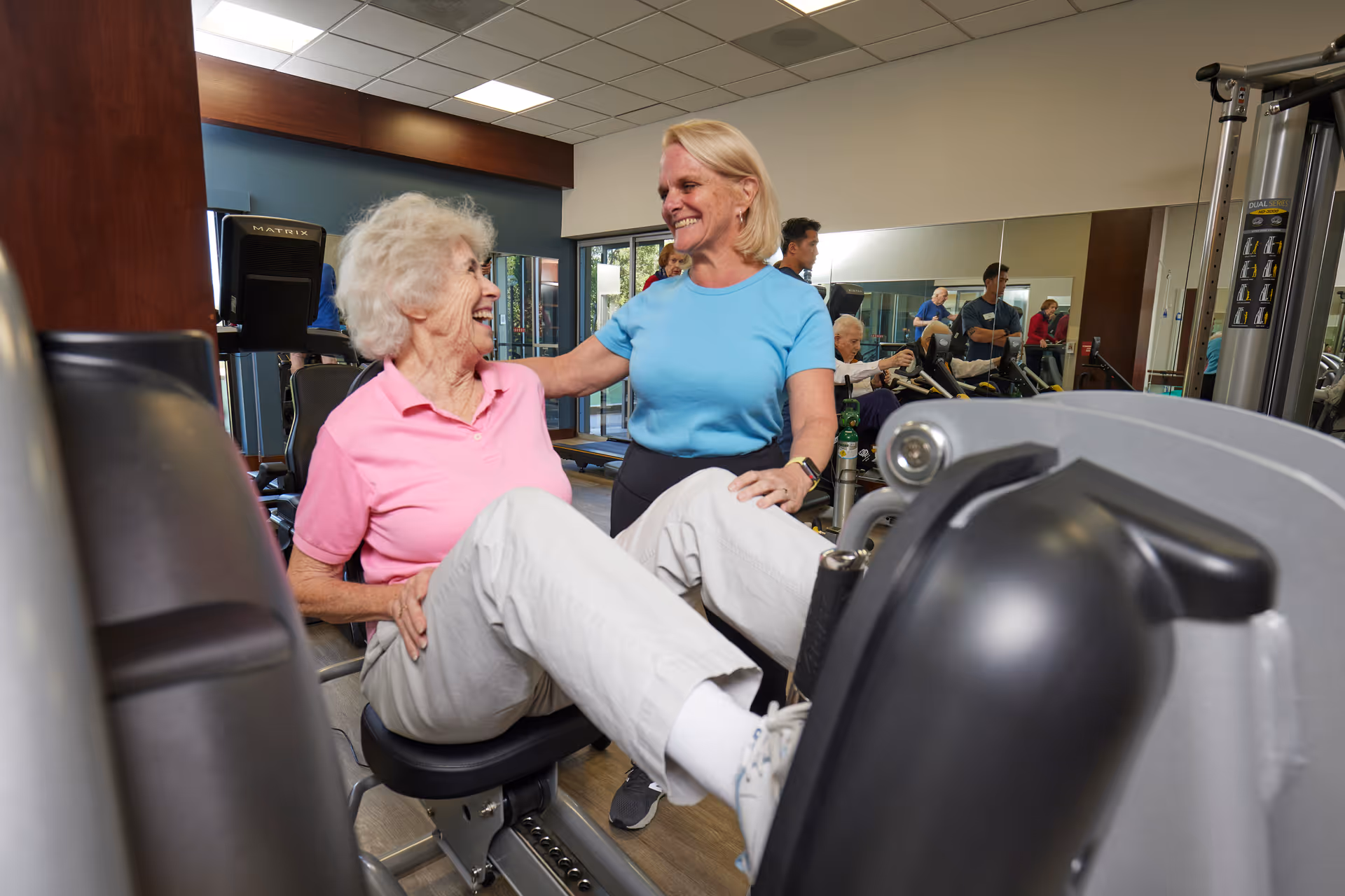 An elderly woman in a pink shirt is exercising on a leg press machine in a gym, assisted by a smiling woman in a blue shirt. Other people are visible in the background using exercise equipment in a well-lit fitness room with mirrors on the wall.