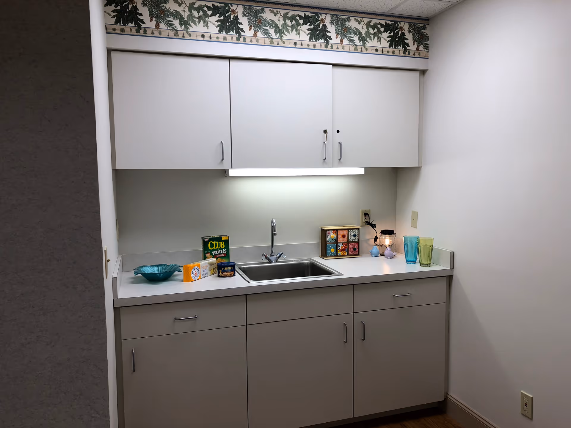 Small kitchenette area with a stainless sink, white upper and lower cabinets, and various items on the countertop.