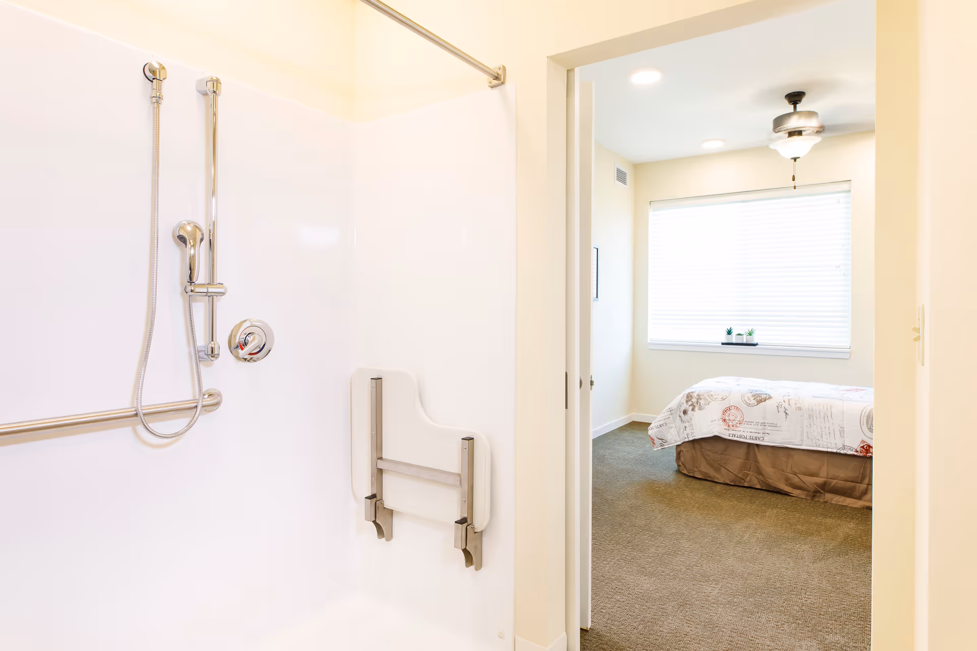 Accessible shower with a handheld showerhead, grab bar and fold-down seat, viewed into a bright bedroom with a bed and window.