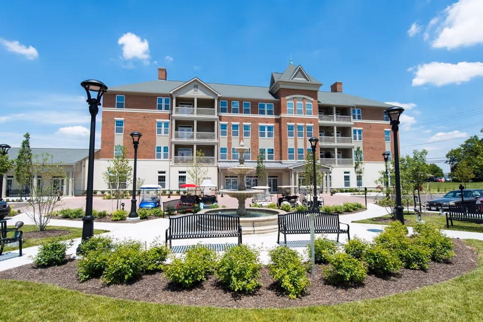 Exterior view of a multi-story senior living facility building with red brick and beige siding. In front of the building is a landscaped garden area with green bushes, a circular fountain, black benches, and several black lamp posts under a blue sky with scattered clouds.