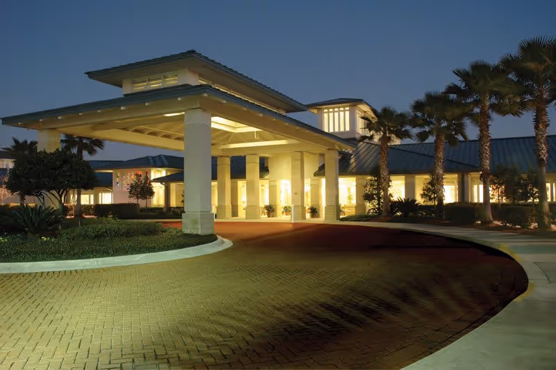 Exterior view of Westminster St. Augustine senior living facility at dusk, showing a covered entrance with bright lighting, palm trees, and landscaped greenery.