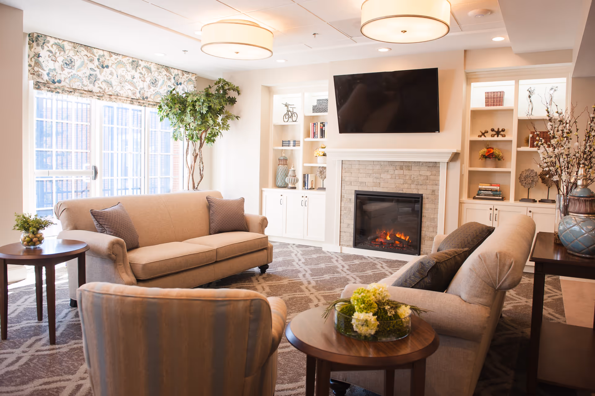 A cozy senior living room with beige sofas and armchairs arranged around a wooden coffee table with a floral centerpiece. The room features a fireplace with a stone surround and a flat-screen TV mounted above it. Built-in white shelves on either side of the fireplace hold decorative items and books. Large windows with patterned valances allow natural light to fill the space, and a tall green plant stands near the window.