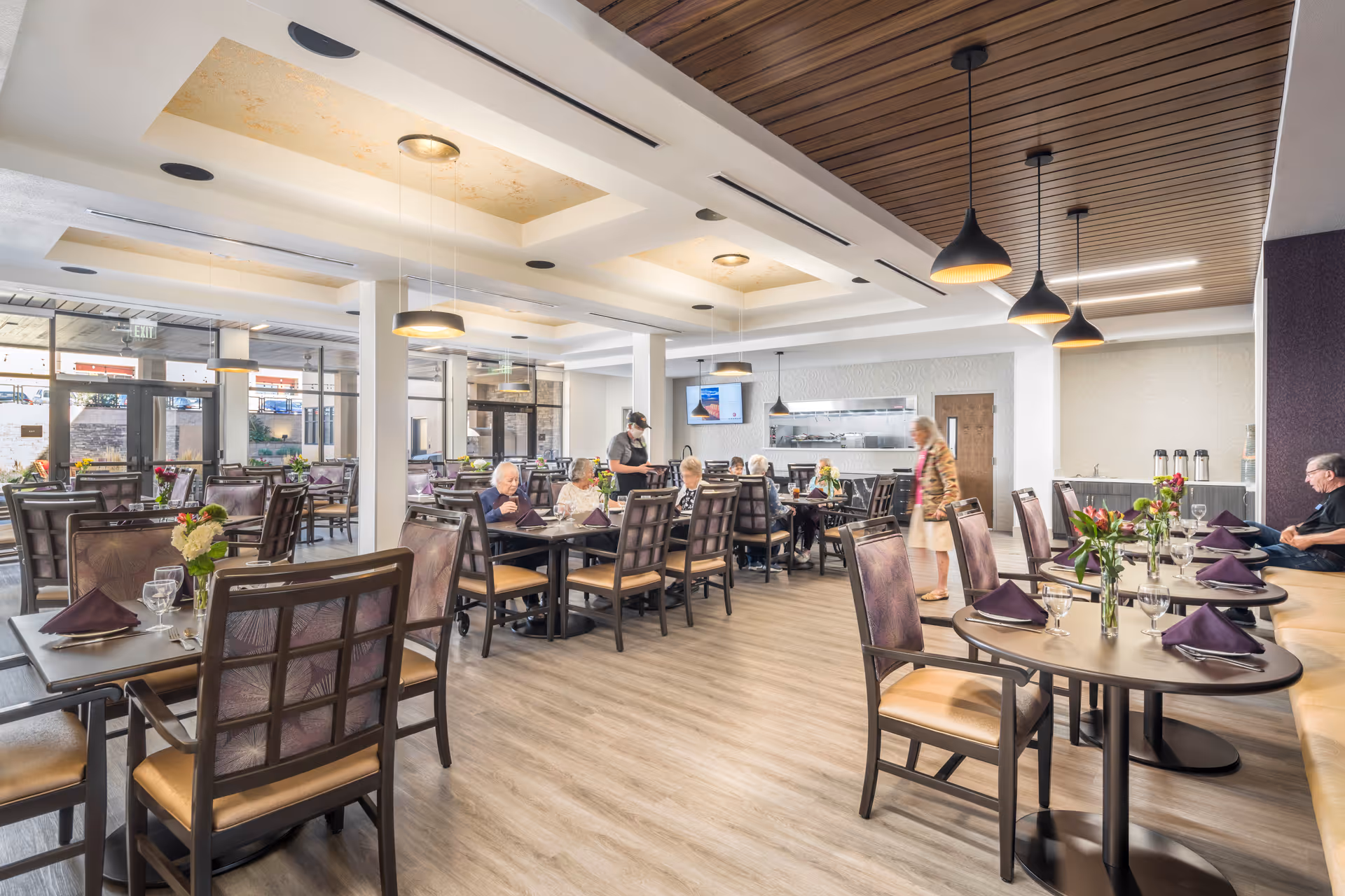 Spacious modern dining room with multiple set tables, pendant lights, and residents and staff interacting.
