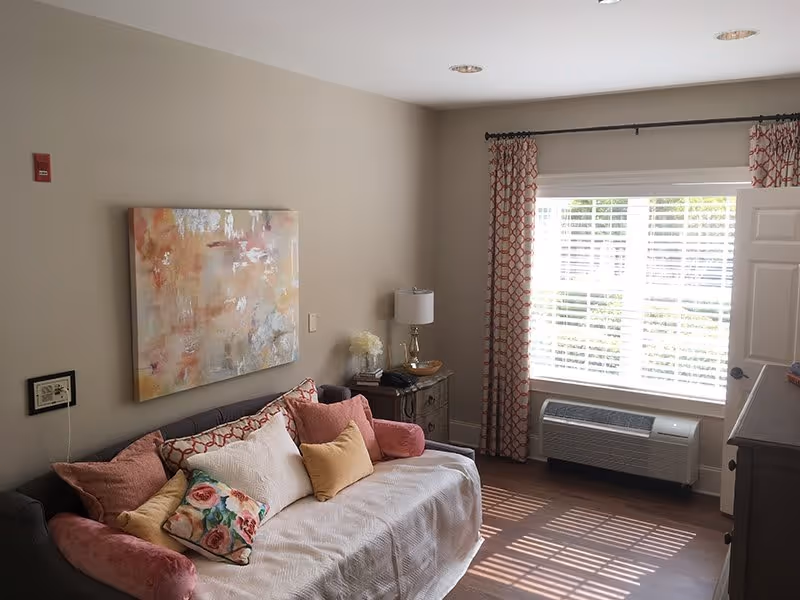 A cozy living room area with a gray sofa covered by a white throw and decorated with multiple colorful pillows including floral and solid colors. Above the sofa hangs an abstract painting with warm tones. To the right of the sofa is a small wooden side table with a lamp, books, and a decorative bowl. A large window with white blinds and patterned red curtains allows natural light to fill the room. Below the window is a wall-mounted air conditioning unit. The room has beige walls and wooden flooring.