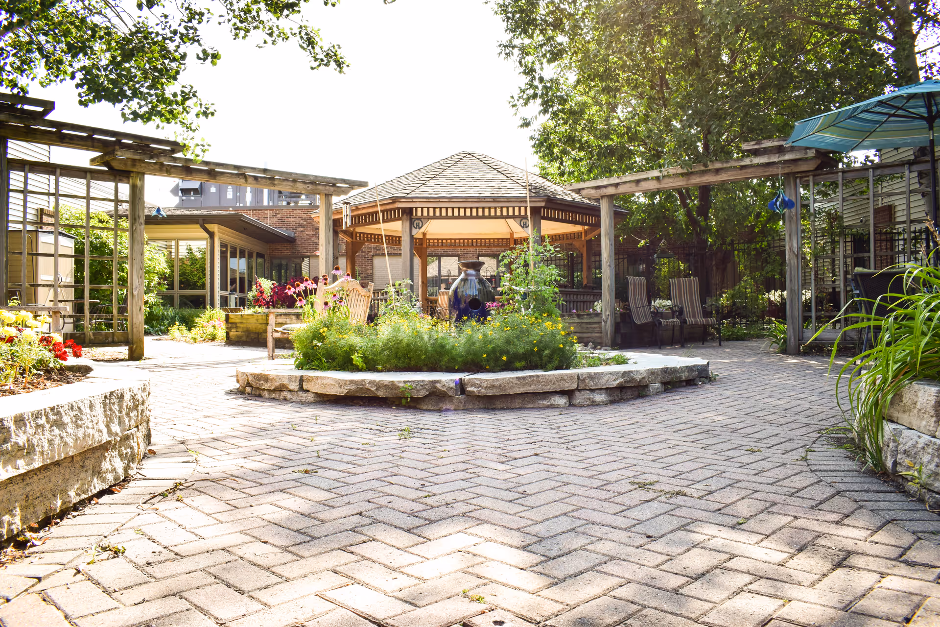Sunny courtyard with a central gazebo, paved brick walkway, raised flower beds, pergolas and outdoor seating.