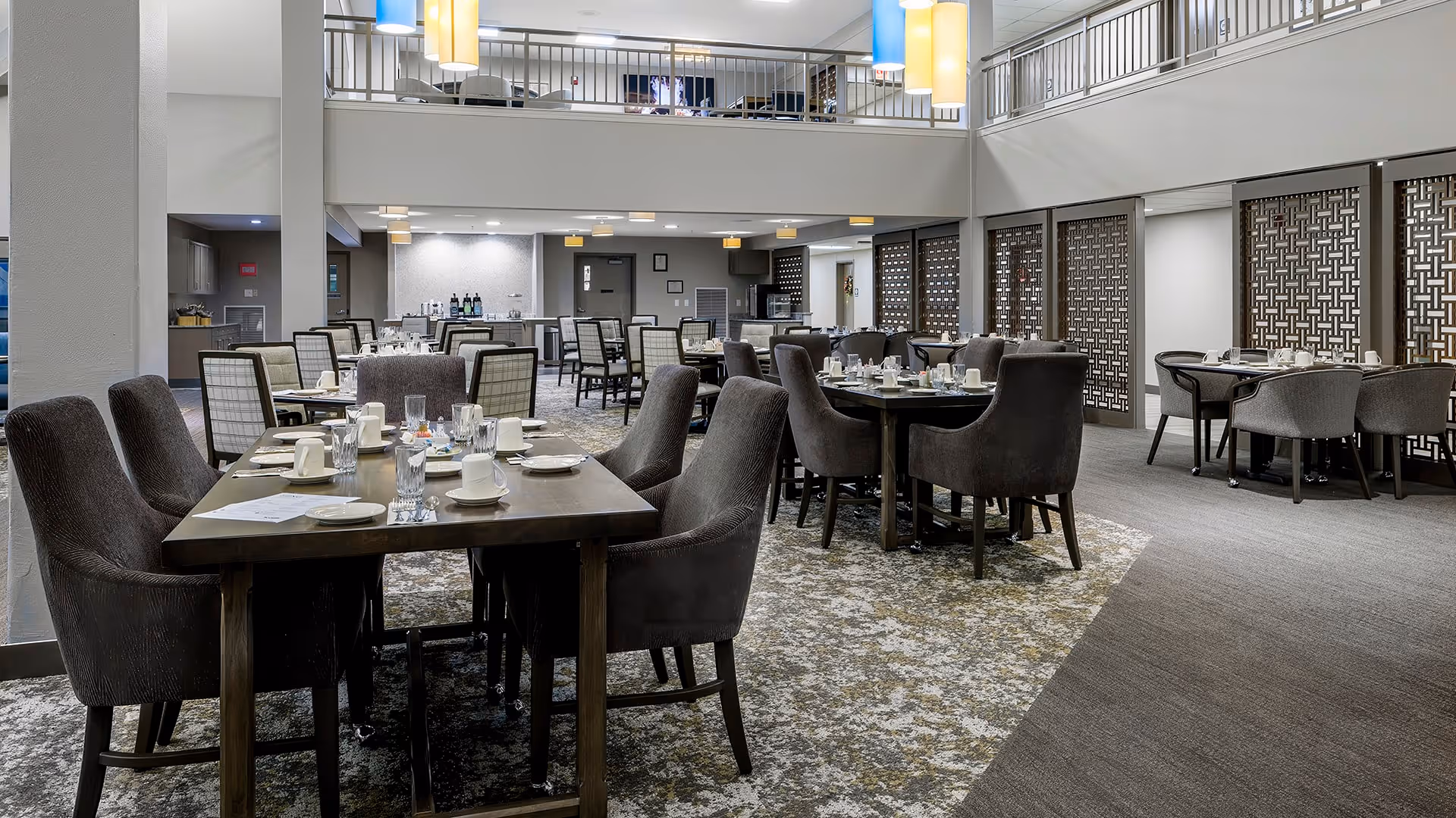 A spacious dining room with multiple tables set with plates, cups, and glasses. The room features comfortable upholstered chairs, patterned carpet, and decorative hanging lights. There is a mezzanine level with a railing overlooking the dining area below.