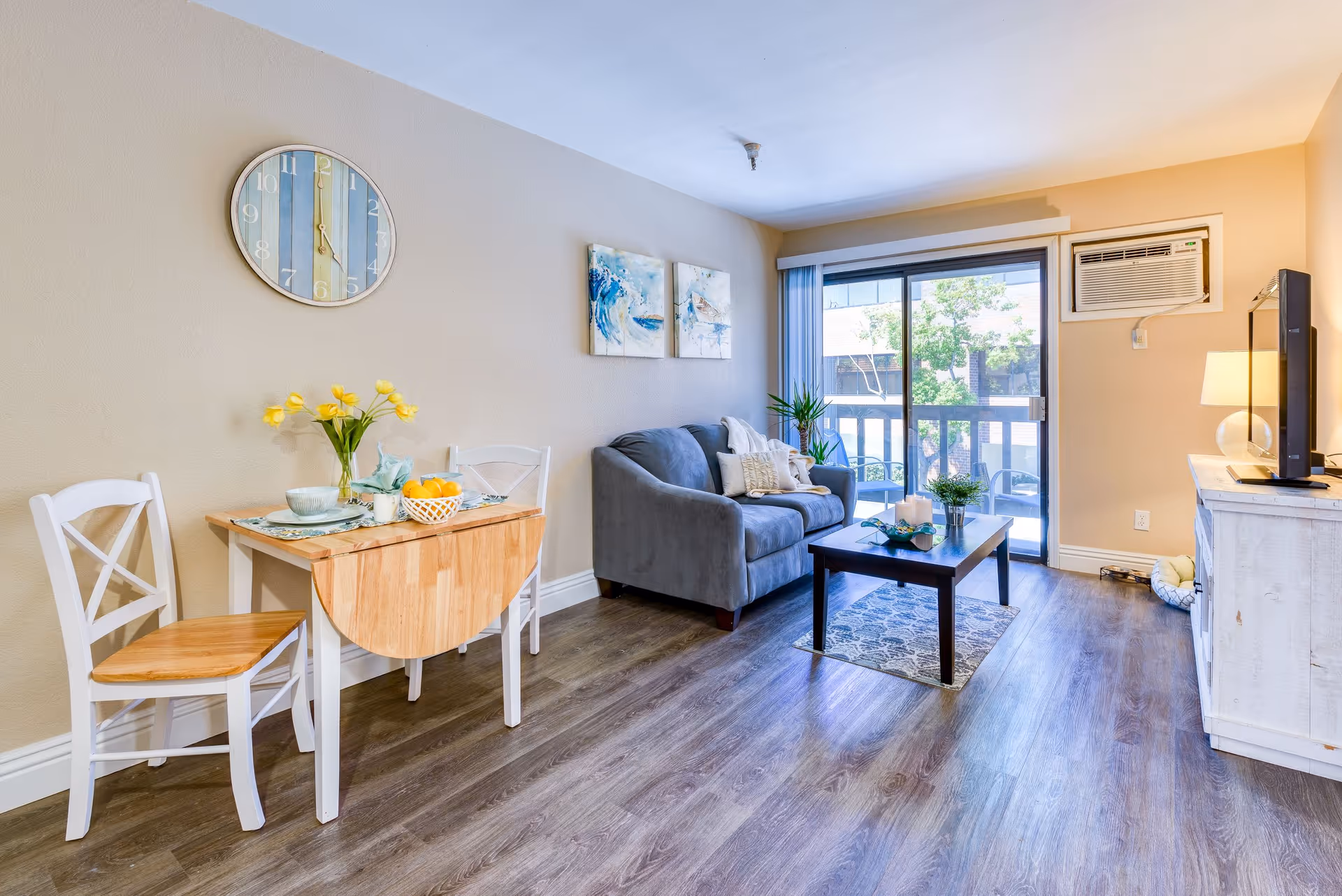 A cozy living room area with a small wooden dining table and two chairs on the left, a gray couch with pillows in the center, a wooden coffee table with decorative items, and a TV on a white wooden stand on the right. There is a large window with vertical blinds and an air conditioning unit above it. The walls are beige, and the floor is wood laminate.