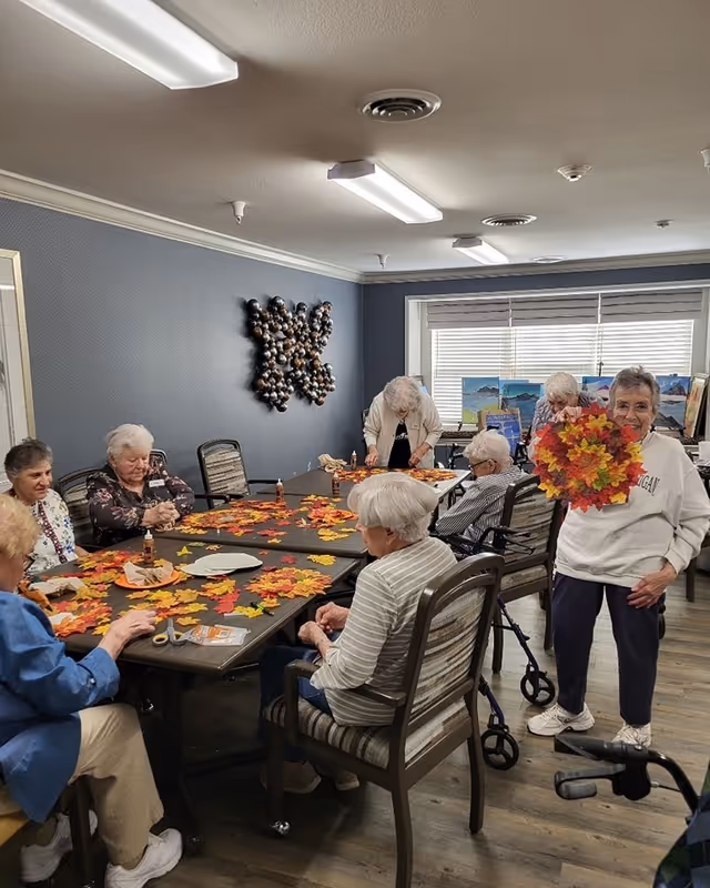 A group of elderly women seated around tables in a room, engaging in a craft activity with colorful autumn leaves. One woman stands holding a circular wreath made of fall leaves, smiling at the camera. The room has blue walls, a decorative wall hanging, and large windows with blinds letting in natural light.