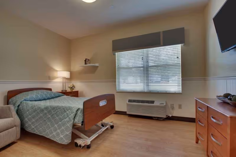 Private senior-care bedroom with a hospital-style bed, armchair, nightstand with lamp, dresser, wall-mounted TV, and a window with blinds.