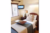 A neatly made single bed in a well-lit room with a wooden headboard, a bedside table with a lamp, a window with blinds, and a wall-mounted flat-screen TV.