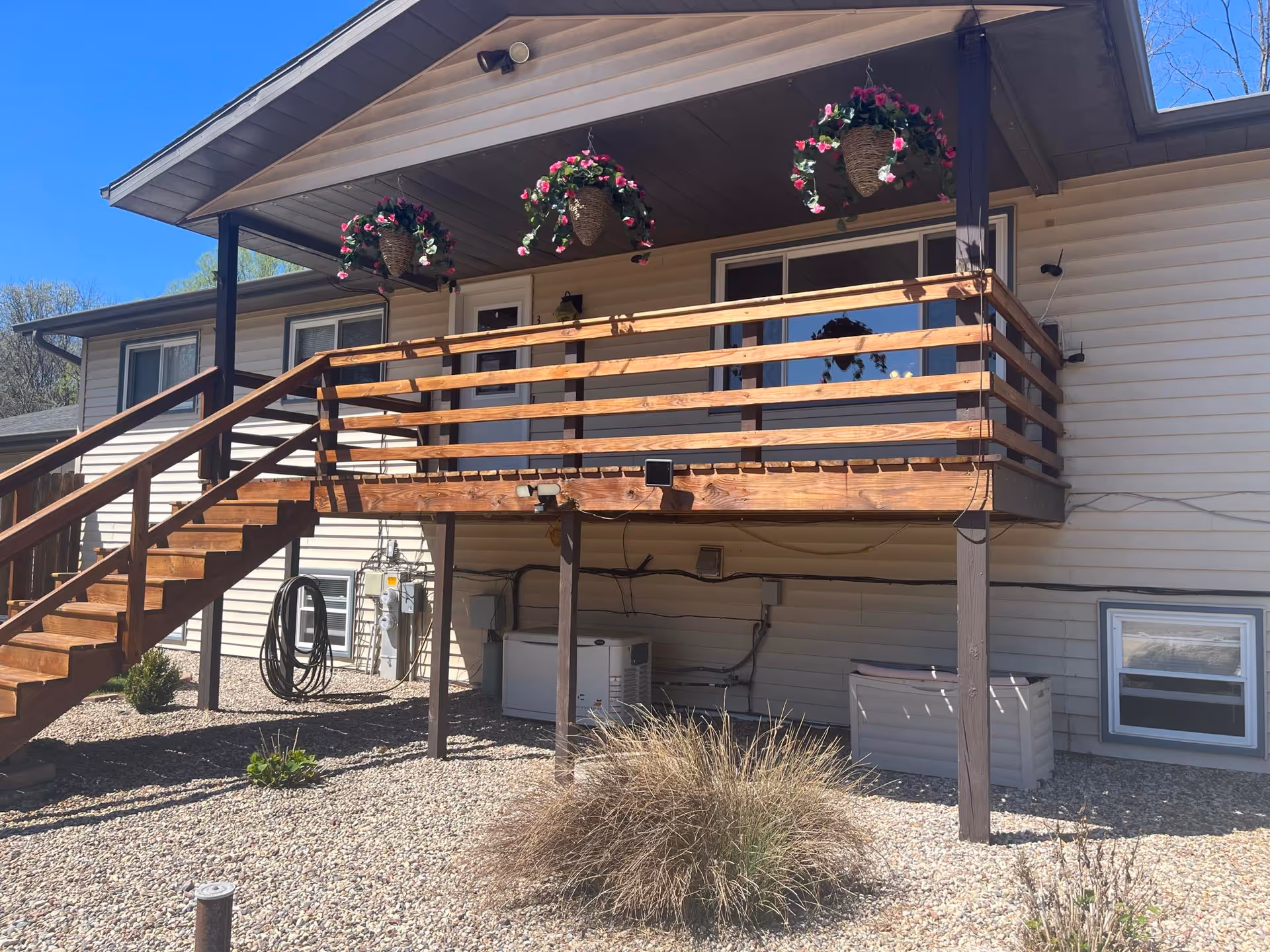 Exterior view of a beige two-story building with a wooden staircase leading to a covered balcony. The balcony has wooden railings and three hanging baskets with pink and white flowers. The ground is covered with small rocks and some plants, and there are utility meters and equipment attached to the building's wall.