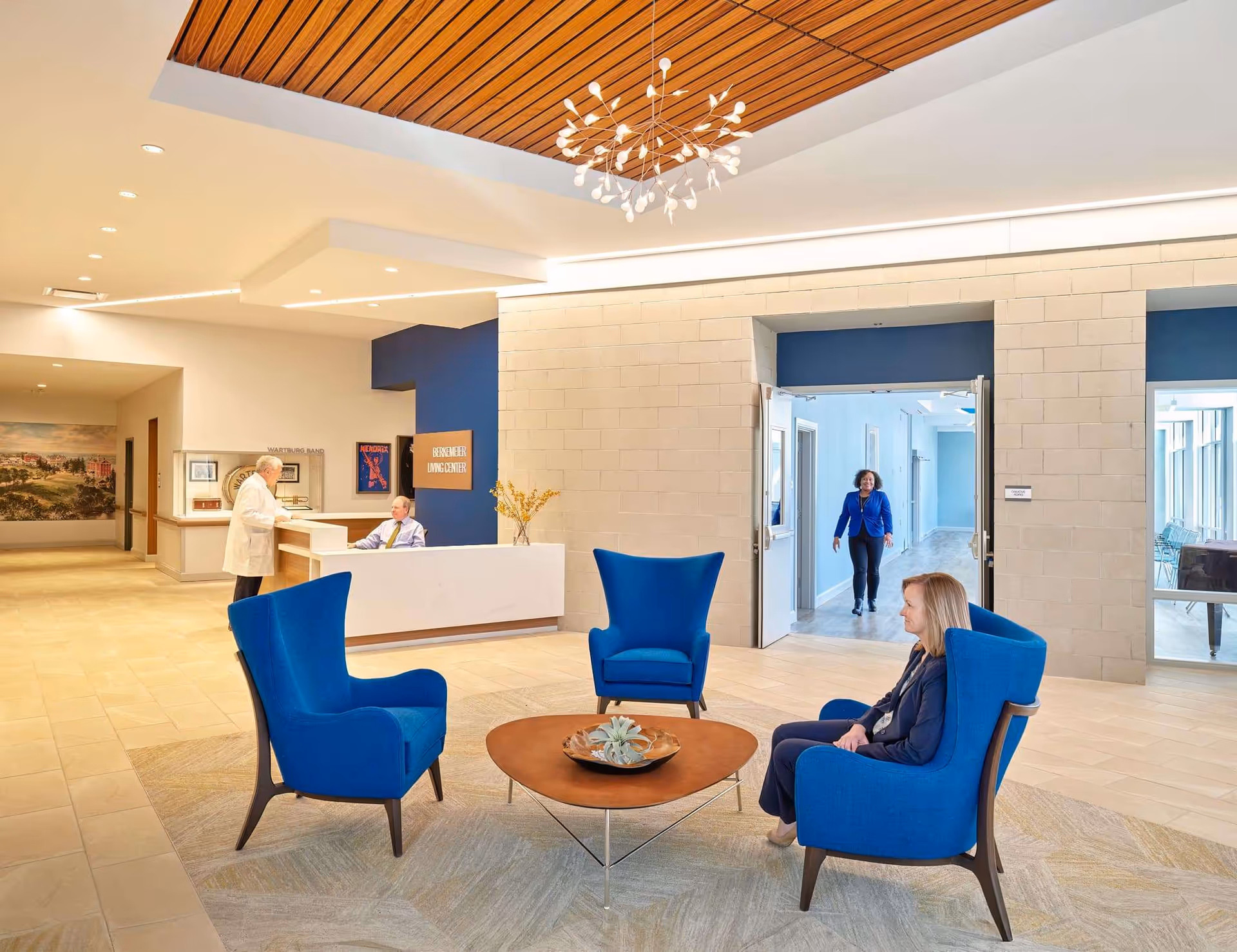 A bright and spacious reception area in The Wartburg Home featuring a modern white reception desk with two staff members behind it. In the foreground, three blue armchairs surround a wooden coffee table with a decorative centerpiece. A woman is seated in one of the chairs, and another woman is walking through double doors in the background. The room has light-colored tile flooring, a wooden ceiling accent, and a contemporary chandelier.