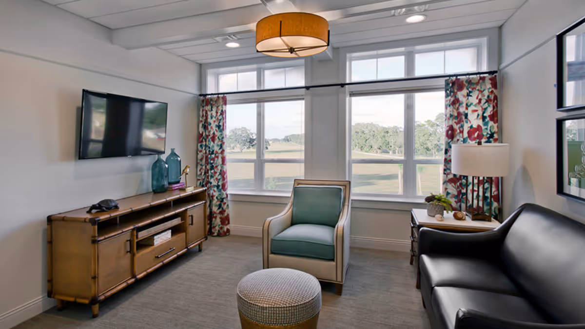 A cozy living room with a large window showing a scenic outdoor view. The room features a wall-mounted flat-screen TV above a wooden console table with decorative items, a cushioned armchair with a blue pillow, a round ottoman, a black leather sofa, a side table with a lamp and small plant, and floral curtains framing the window.