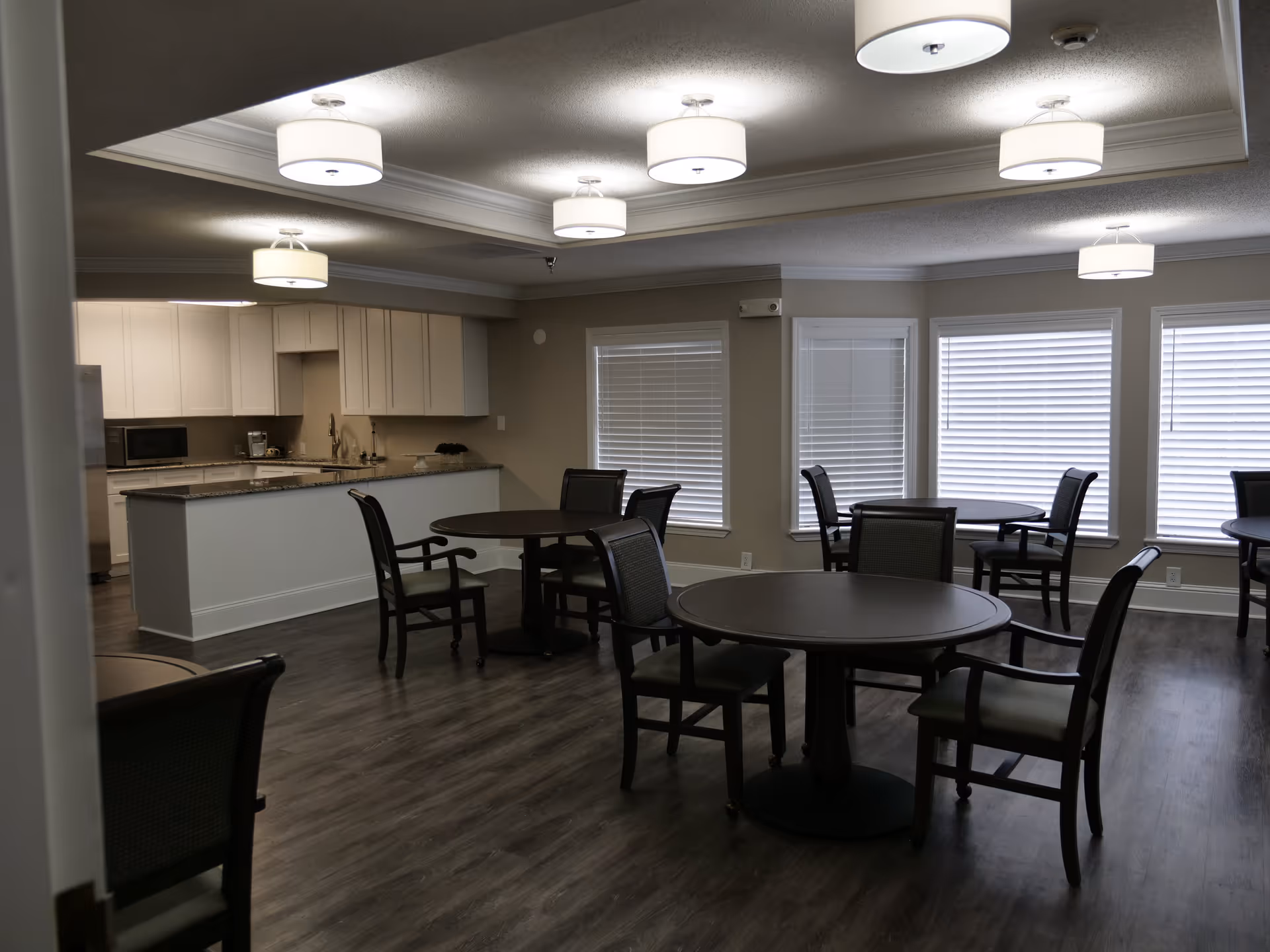Interior view of a dining area in a senior living facility with round tables and chairs arranged on a wood floor. The room has multiple windows with blinds and ceiling lights. A kitchen area with white cabinets and a granite countertop is visible in the background.