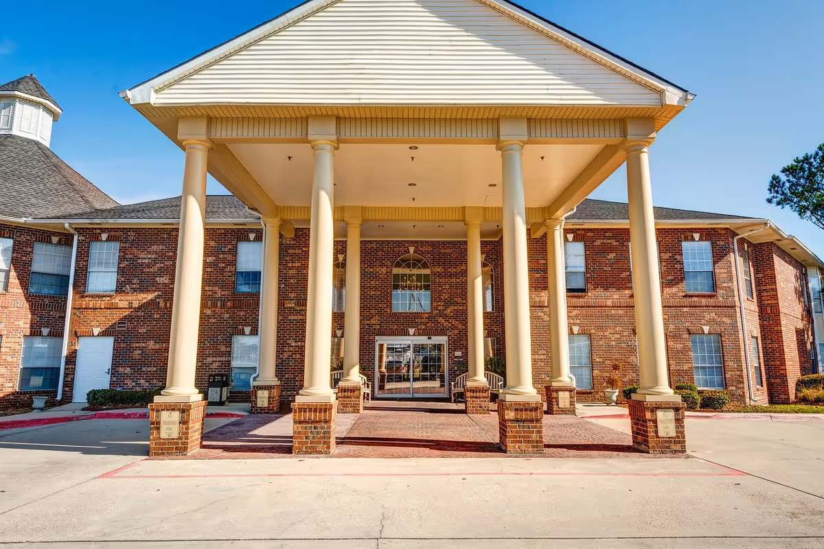 Front exterior view of a senior living facility named Laketown Village, featuring a large covered entrance supported by six tall white columns, red brick walls, multiple windows, and a clear blue sky.