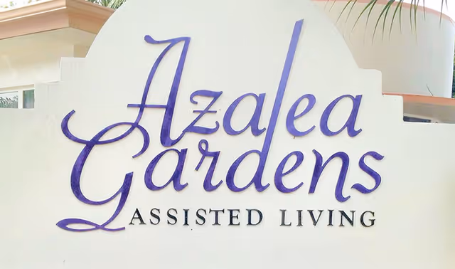 Sign for Azalea Gardens Assisted Living facility with purple cursive lettering on a white background, part of a building and some palm leaves visible behind the sign.