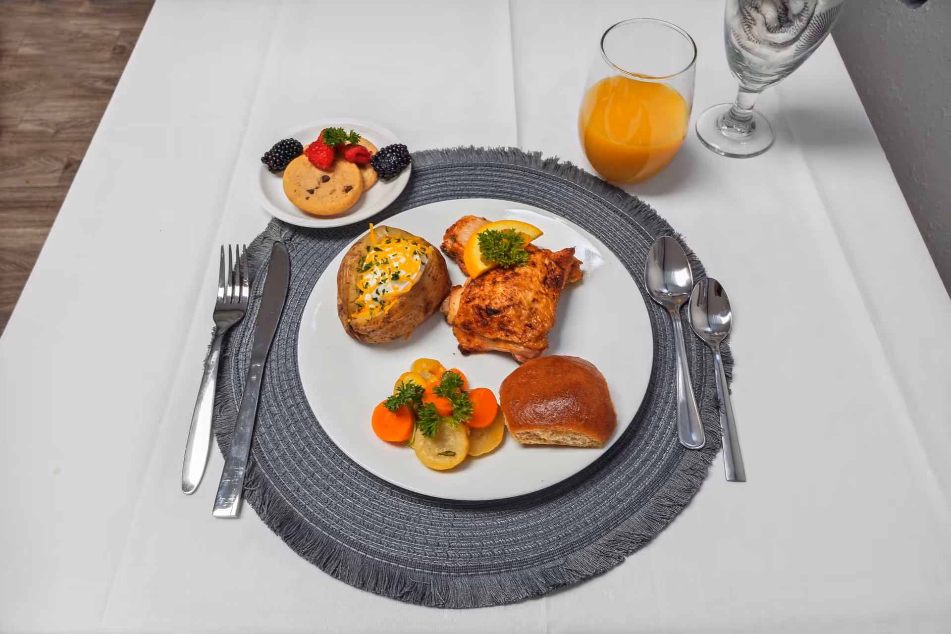 A neatly arranged meal on a white plate with a baked potato topped with cheese and sour cream, a piece of grilled chicken garnished with a lemon slice and parsley, cooked vegetables, and a bread roll. Next to the plate is a small dish with a chocolate chip cookie and fresh berries. There is a glass of orange juice and a glass of water on the white tablecloth, with silverware placed on either side of the plate on a gray woven placemat.