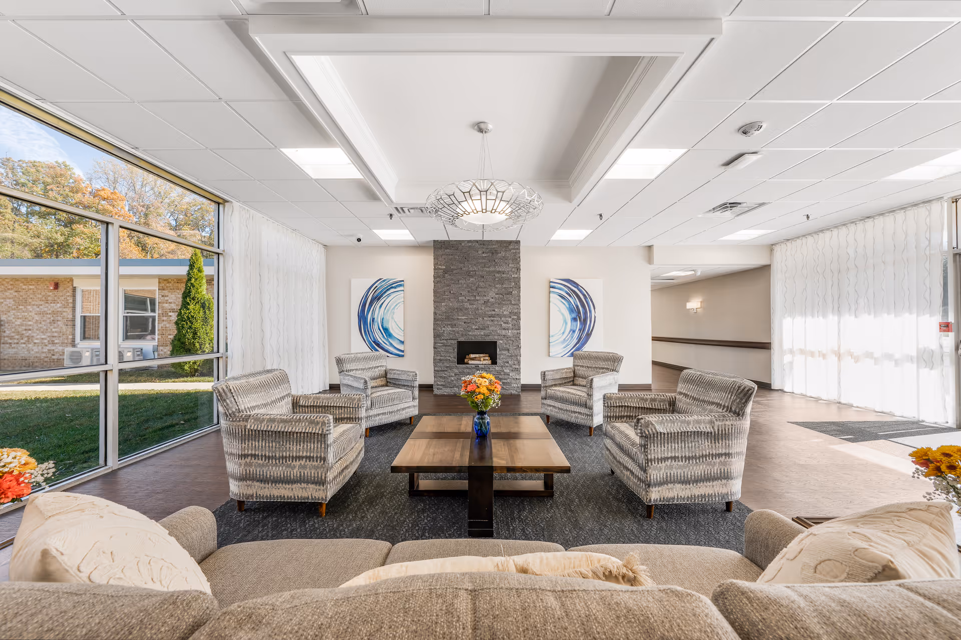 Bright communal lounge with sofas and armchairs arranged around a central coffee table and a stone fireplace, large windows letting in natural light.