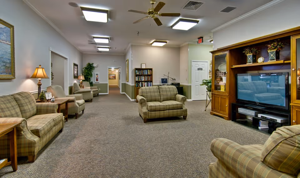 A spacious senior living facility common area with plaid upholstered armchairs and loveseats arranged around a wooden entertainment center with a flat-screen TV. The room has carpeted floors, soft lighting from ceiling fixtures and table lamps, framed artwork on the walls, and a bookshelf with books and a computer in the background.