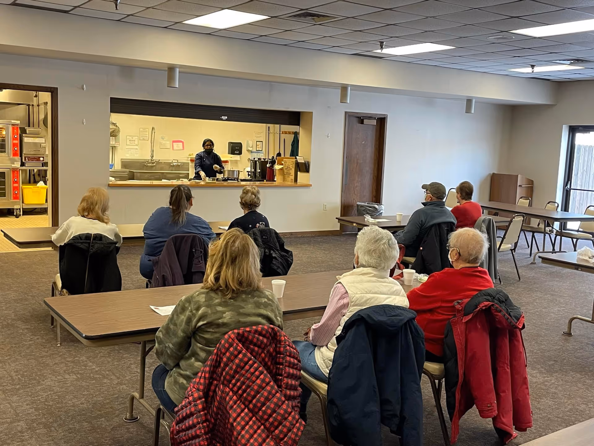 A group of elderly people seated at tables in a community room, facing a kitchen pass-through window where a staff member is preparing food. The room has a carpeted floor, a drop ceiling with fluorescent lights, and several empty tables and chairs along the walls.