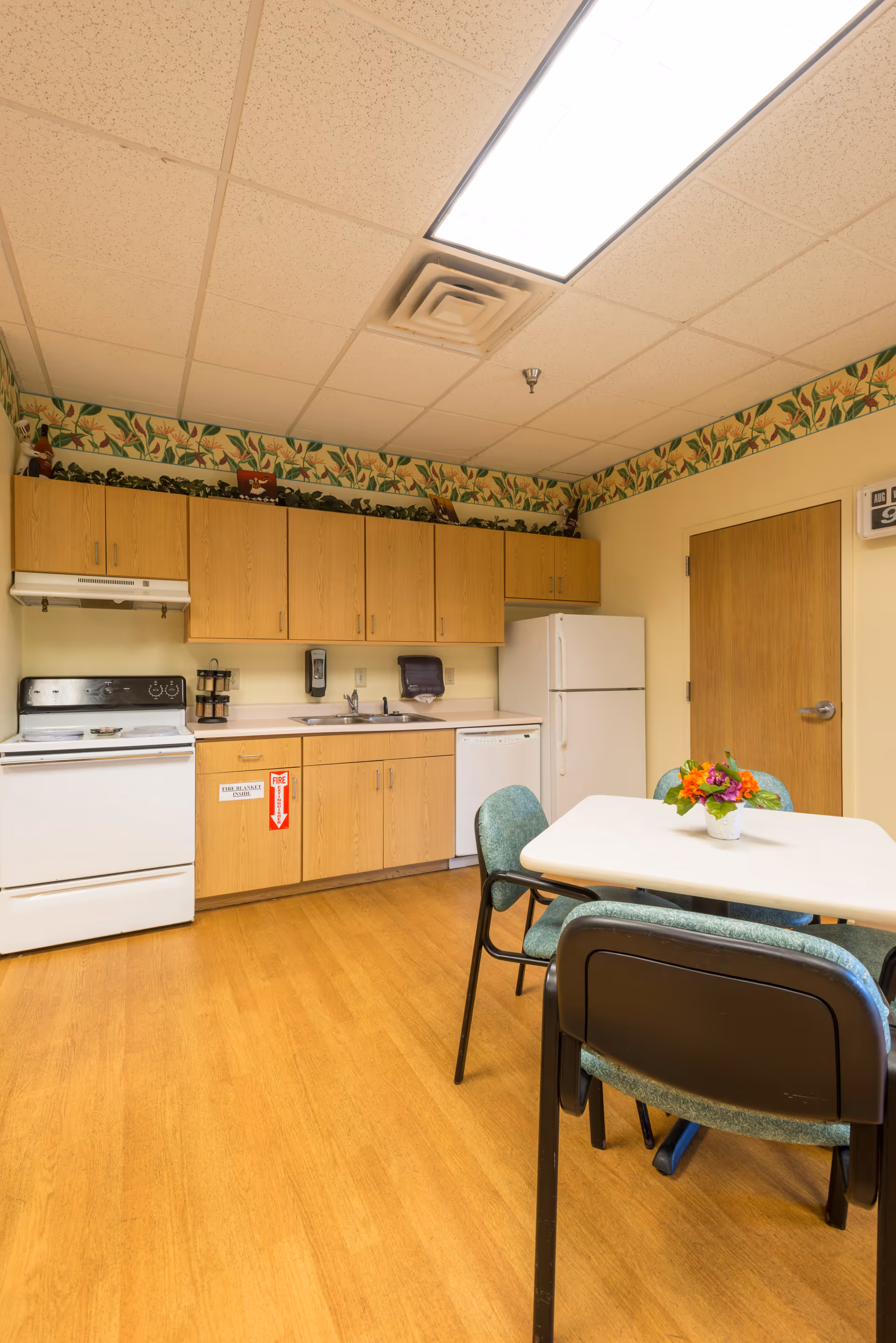 A small kitchen area in a skilled nursing facility with wooden cabinets, a white stove, refrigerator, and dishwasher. There is a white table with four green cushioned chairs around it, and a small flower arrangement on the table. The walls have a floral border near the ceiling, and the floor is wood laminate.