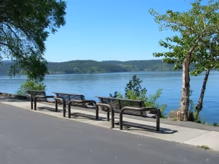 A paved walkway along a body of water with several empty benches facing the water. Trees and greenery are visible along the edge of the walkway, and hills can be seen in the distance across the water under a clear blue sky.