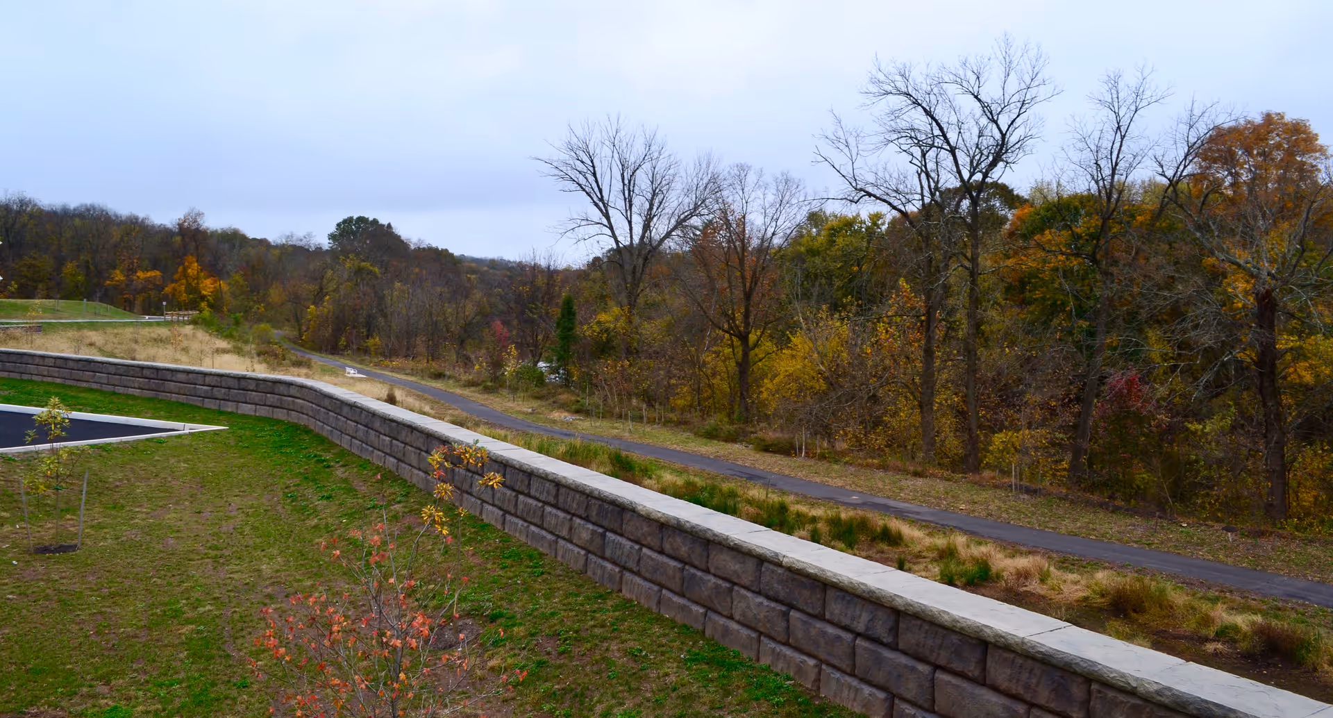 View over a stone retaining wall toward a paved path and wooded area with autumn foliage under an overcast sky.